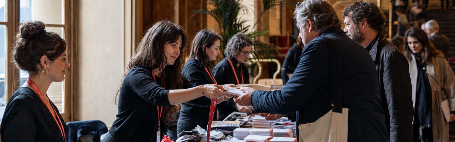 Freiwillige geben am Akkreditierungsschalter des Festivals gedruckte Badges und Programmhefte aus.